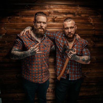 Two mans with beards and tattoo posing over wooden wall. One of them holding axe.