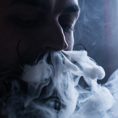 Man with Beard and Mustages Vaping an Electronic Cigarette. Vaper Hipster Smoke Vaporizer and Exhals Smoke Flow from Nose.