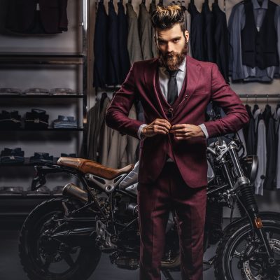 Handsome man with a stylish beard and hair dressed in vintage red suit posing near retro sports motorbike at men's clothing store.